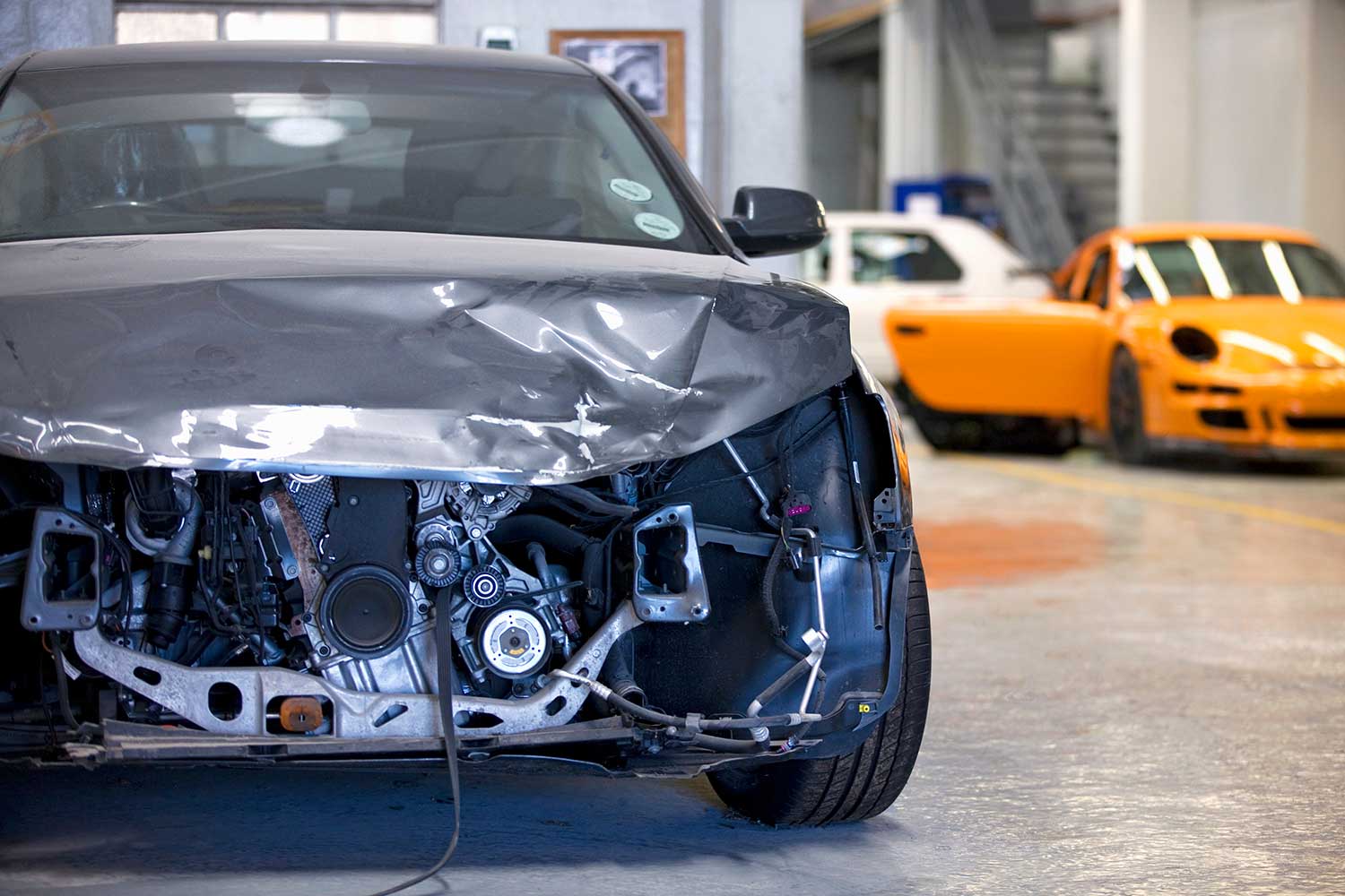 A gray car with a severely crumpled hood and exposed engine sits in an auto repair shop; a bright orange sports car is parked behind it, contrasting the damage.