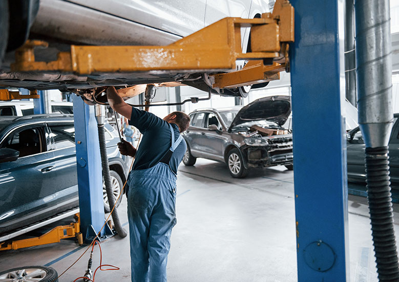 A mechanic in blue overalls works under a car on a lift in a busy auto repair shop, with several vehicles in the background.