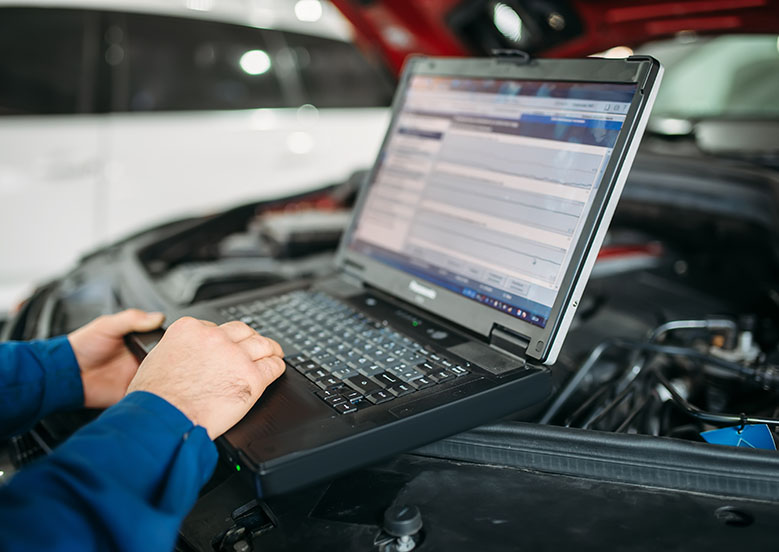 A mechanic analyzes vehicle diagnostics on a laptop placed on a car's engine, in a bright service garage.