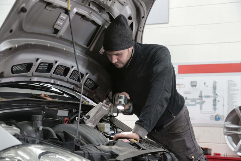 A mechanic in a black outfit examines a car engine while holding a flashlight, surrounded by garage equipment and manuals.